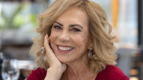 A personagem Frida Mancini (Arlete Salles) sorridente, com a mão no rosto e vestindo uma roupa vermelha