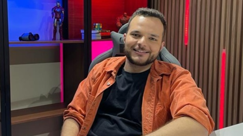 Leonardo Zalcman, apresentador da PlayTV, sentado em uma cadeira gamer e com o braço apoiado em uma mesa branca