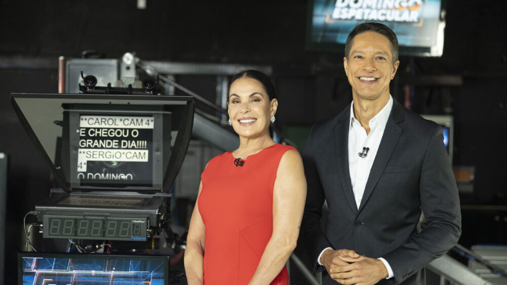 Carolina Ferraz e Sergio Aguiar nos bastidores do Domingo Espetacular, em frente a uma câmera que mostra o texto que será lido pelos apresentadores