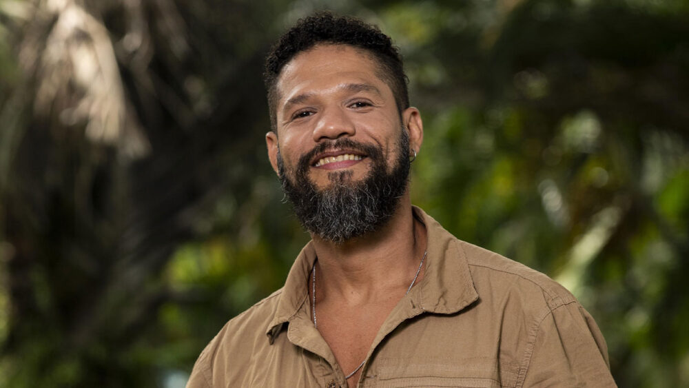 Juninho, ex-participante do BBB 24, sorridente em foto no jardim dos Estúdios Globo