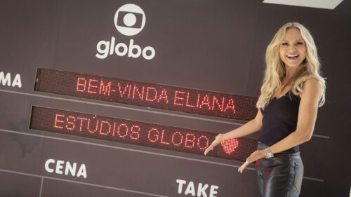 Eliana Michaelichen durante sua visita aos Estúdios Globo. Ela está na frente de uma claquete gigante em que aparece escrito no painel digital "bem-vinda Eliana"