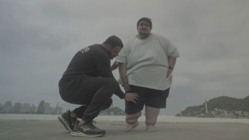 Imagem com foto de participante de Quilos Morais Brasil na praia com profissional da saúde ao lado dele agachado na areia