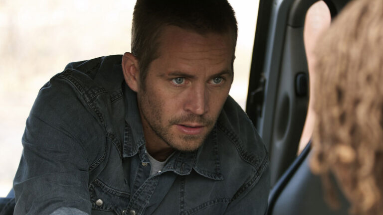 Ator Paul Walker com cabelo curto, barba e camisa jeans olhando atentamente para alguém em cena de Veículo 19