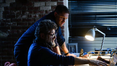 Tom Hardy e Andy Serkis em cena de Venom Tempo de Carnificina olhando para computador em ambiente escuro iluminado por luminária