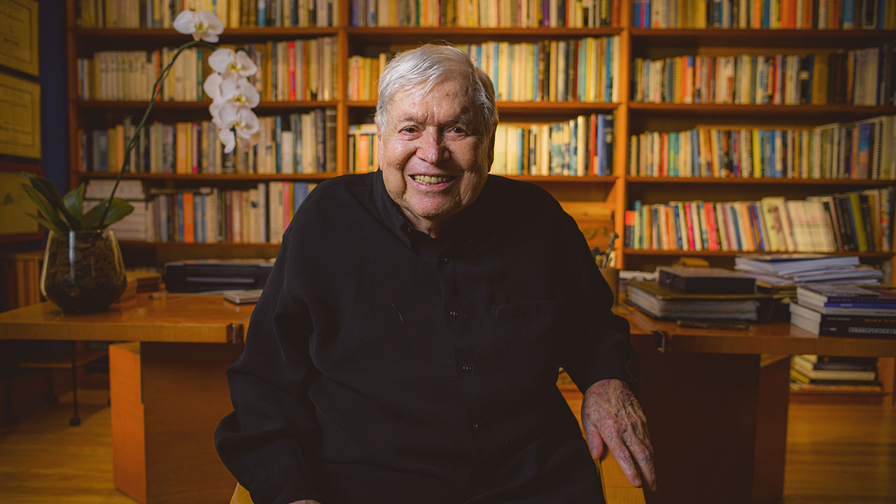 José Bonifácio de Oliveira Sobrinho, o Boni, sorridente com biblioteca ao fundo durante o documentário Tributo