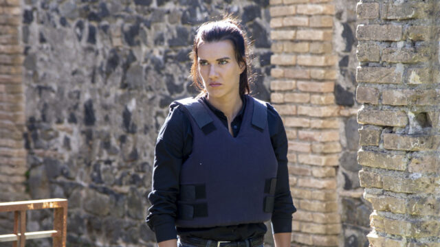 Natalie Burn como Emma usando colete à prova de balas e expressão séria caminha entre ruínas de pedra no filme Sobreviva à Noite (2019)