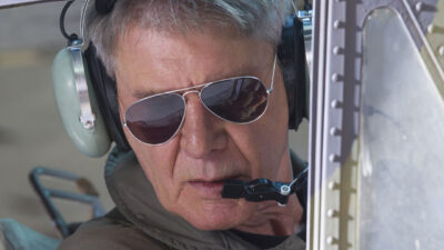 Harrison Ford como Max Drummer usando óculos escuros e fones de piloto em cena do filme Os Mercenários 3