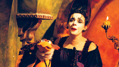 Rosi Campos como Morgana e a ave Adelaide em cena de Castelo Rá-Tim-Bum, com cenário iluminado em tons quentes ao fundo