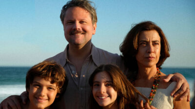 Elenco do filme Ainda Estou Aqui posa na praia ao pôr do sol, com os personagens demonstrando expressões contrastantes