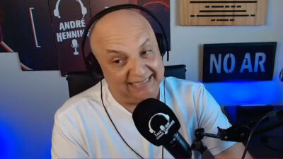 André Henning usa fones de ouvido e camiseta branca enquanto fala ao microfone em estúdio com letreiro "no ar" ao fundo