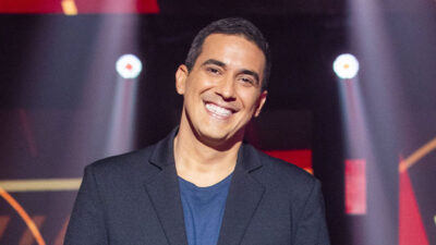 André Marques sorri no palco do The Voice+, da Globo, vestindo blazer preto e camisa azul, com luzes de palco ao fundo