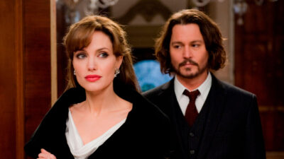 Angelina Jolie e Johnny Depp em cena de O Turista, filme da Band. Ela está vestindo casaco preto e ele de terno escuro, em ambiente sofisticado
