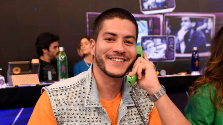 Arthur Aguiar sorri enquanto atende um telefone verde no Criança Esperança. Ele veste colete jeans com tachinhas sobre camiseta laranja