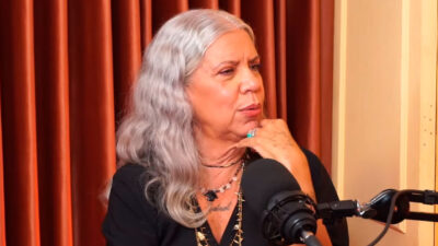 Astrid Fontenelle durante entrevista em um podcast, vestindo roupa preta, com microfone à frente e cortina avermelhada ao fundo