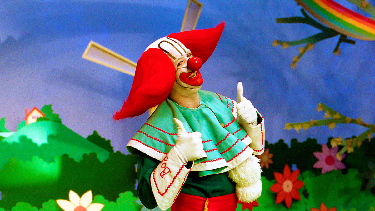 Bozo posa sorridente no cenário colorido de seu programa infantil no SBT, usando figurino tradicional com peruca vermelha e nariz de palhaço