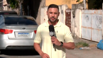 Repórter Bruno Assunção, do SBT Rio, segura um microfone com o logotipo da emissora enquanto grava uma reportagem em uma rua com casas e um carro estacionado ao fundo