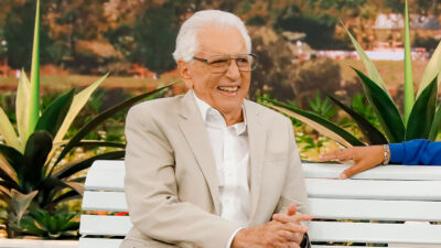 Carlos Alberto de Nóbrega sorri enquanto está sentado em um banco branco, usando terno bege, com plantas e cenário natural ao fundo