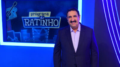 Carlos Massa, o Ratinho, sorri no cenário do Programa do Ratinho, com o logotipo do programa iluminado ao fundo