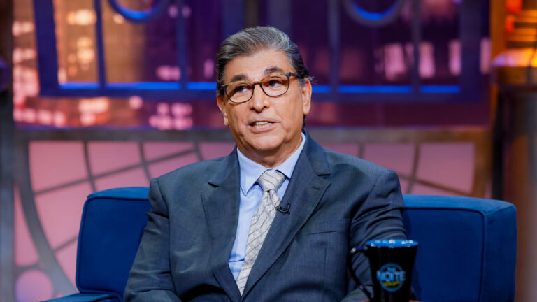 Carlos Nascimento durante entrevista no The Noite, sentado em um sofá azul, vestindo terno e gravata, com o cenário iluminado ao fundo
