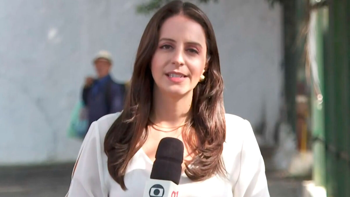 Carol Ianelli faz uma reportagem ao vivo segurando um microfone da TV Globo, vestindo blusa clara, com fundo desfocado ao ar livre