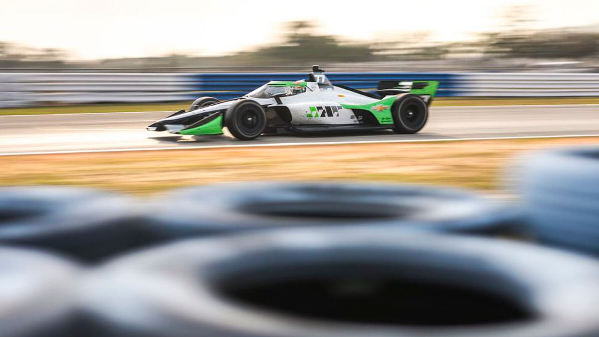 Carro de Fórmula Indy em alta velocidade na pista, com pintura preta, branca e verde, e barreira de pneus desfocada no primeiro plano
