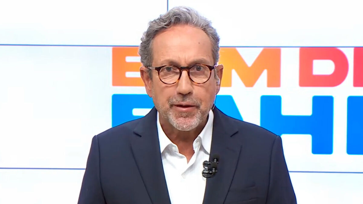 Apresentador Casemiro Neto no estúdio do programa Bom Dia Bahia, vestindo terno escuro e óculos, com expressão séria