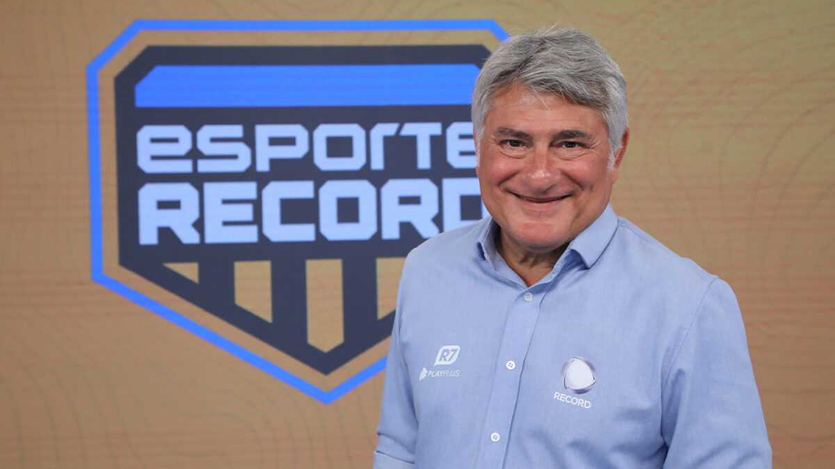 Cléber Machado sorri em frente ao logo do programa Esporte Record, vestindo camisa azul com o emblema da Record