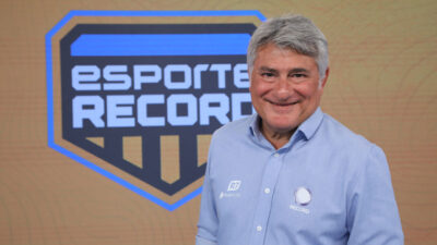 Cléber Machado sorri em frente ao logo do programa Esporte Record, vestindo camisa azul com o emblema da Record