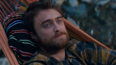 Yossi Ghinsberg (Daniel Radcliffe) com barba e camisa xadrez amarela deitado em uma rede colorida em cena do filme Na Selva