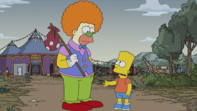 Personagens Krusty e Bart Simpson conversam em um cenário de circo com tendas, árvores e uma girafa ao fundo. Krusty veste roupas coloridas e segura uma trouxa amarrada a um bastão