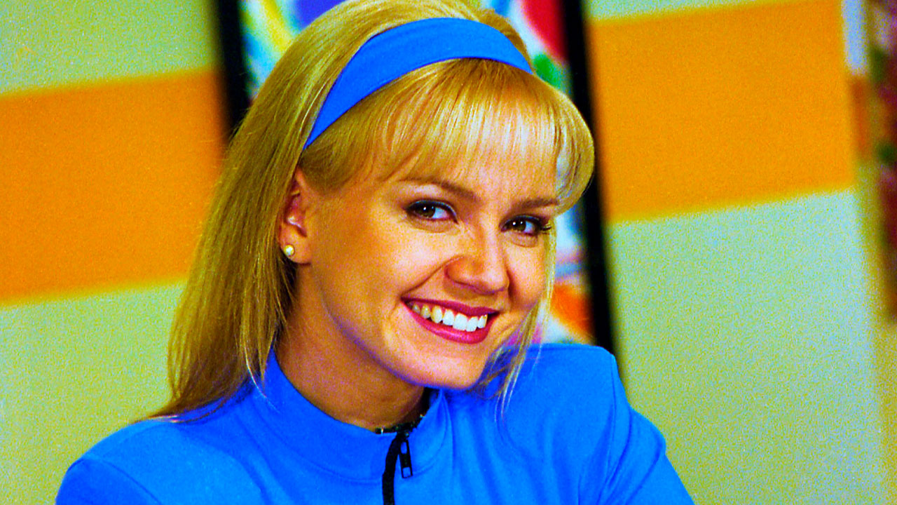 Eliana sorrindo no cenário do programa Eliana & Cia, vestindo roupa azul combinando com a faixa no cabelo