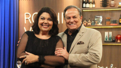 Fabiana Karla e Ronnie Von posam sorridentes no cenário do programa Companhia Certa. Ela veste um vestido preto com mangas de tule, e ele usa um blazer bege com camisa preta. Ao fundo, prateleiras decoradas e um letreiro iluminado
