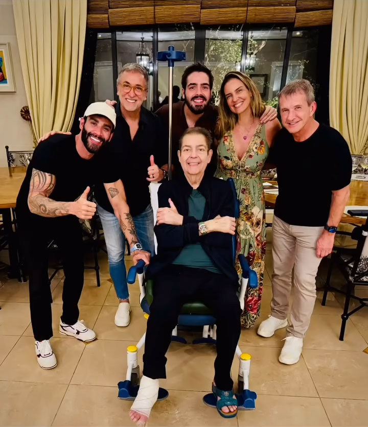 Faustão sentado em uma cadeira de rodas com o pé enfaixado cercado por familiares e amigos sorridentes em um ambiente interno