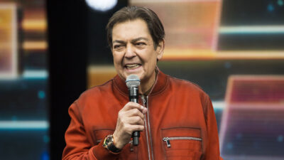 Faustão no palco segurando um microfone e usando jaqueta vermelha enquanto fala ao público