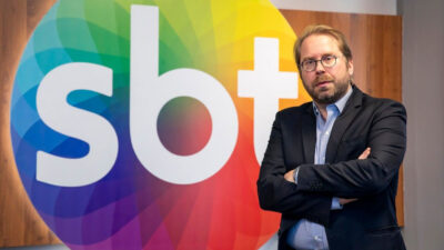 Fernando Justus Fischer posa de braços cruzados em frente ao logotipo colorido do SBT em um ambiente corporativo