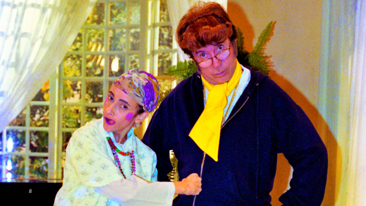 Filó e Moacyr Franco em cena humorística, com Filó vestindo roupa simples e lenço na cabeça e Moacyr de óculos e cachecol amarelo