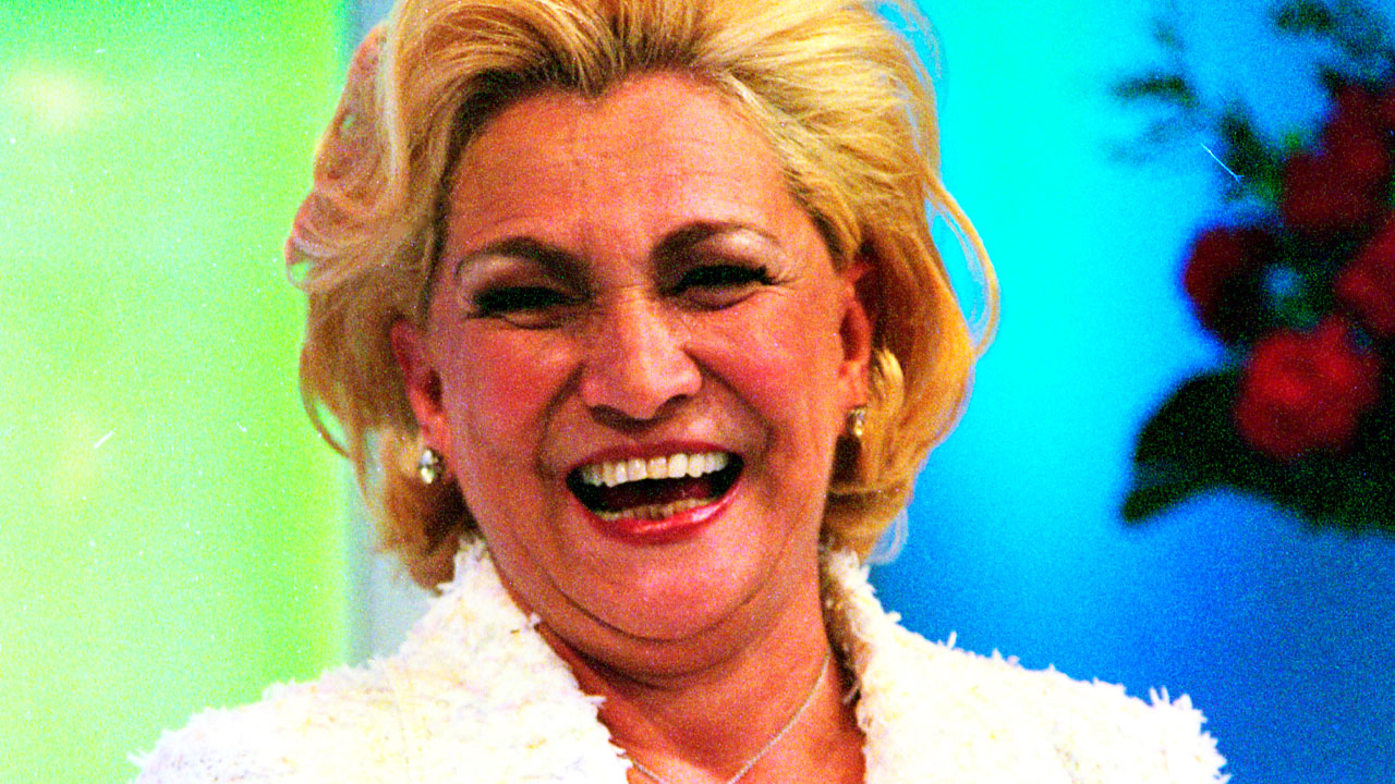 Hebe Camargo sorri no palco de seu programa, vestindo roupa branca e acessórios brilhantes, com fundo colorido e arranjo de flores