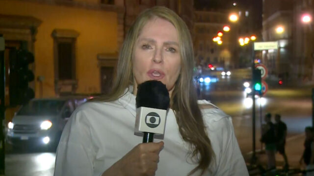 Ilze Scamparini em entrada ao vivo para a TV Globo segurando microfone da emissora com uma cidade iluminada ao fundo