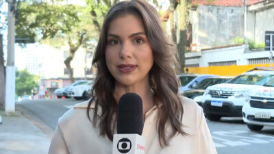 Isadora Markus da TV Globo em reportagem de rua segurando microfone da emissora com viaturas policiais ao fundo