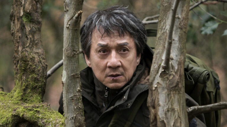 Jackie Chan caracterizado como Quan Ngoc Minh em O Estrangeiro (2017), escondido entre árvores na floresta, usando jaqueta escura e mochila militar