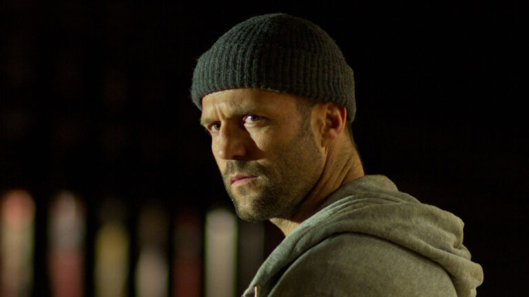 Jason Statham com expressão séria, usando gorro preto e moletom cinza, em cena do filme O Código (2012)