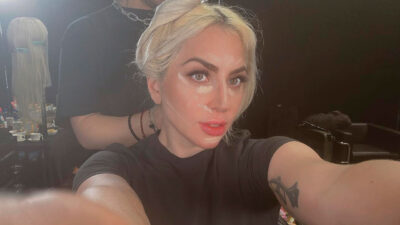 Lady Gaga tira uma selfie durante preparação de maquiagem, com cabelo loiro preso e marcas de contorno no rosto