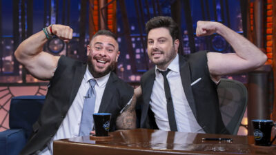 Leo Stronda e Danilo Gentili posam no cenário do programa The Noite com os braços flexionados e mangas do paletó rasgadas