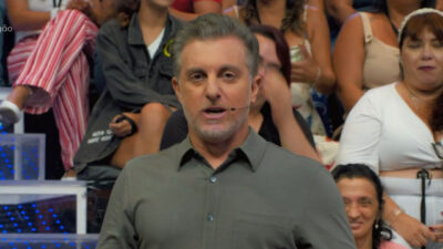 Luciano Huck apresenta seu programa em um estúdio com plateia ao fundo, usando camisa verde e microfone de headset