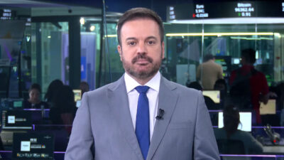 Marcelo Torres veste terno cinza e gravata azul no jornal Real Time, do Times Brasil, com redação e telas de cotações ao fundo