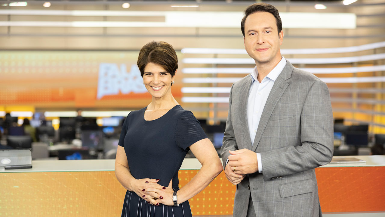 Mariana Godoy e Eduardo Ribeiro sorriem no estúdio do Fala Brasil, com o telão laranja e redação ao fundo