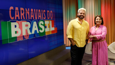 Murilo Ribeiro e Bárbara Pereira posam sorrindo no estúdio do programa Carnavais do Brasil, da TV Brasil, com painel colorido ao fundo