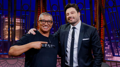Pedro Manso e Danilo Gentili no The Noite, sorrindo no estúdio com fundo iluminado e mesa com caneca do programa