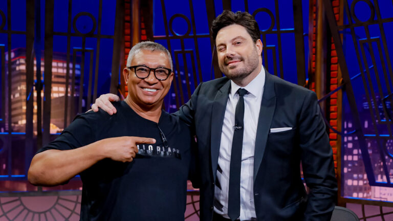 Pedro Manso e Danilo Gentili no The Noite, sorrindo no estúdio com fundo iluminado e mesa com caneca do programa