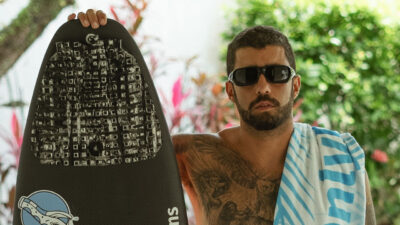 Pedro Scooby ao lado de uma prancha de surfe. Ele está sem camisa e de óculos escuro